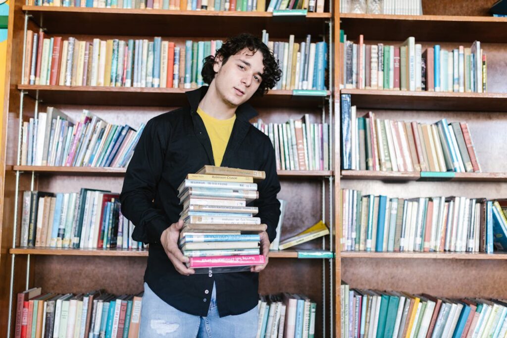 A young adult male holding a stack of books in a library, symbolizing education and learning.